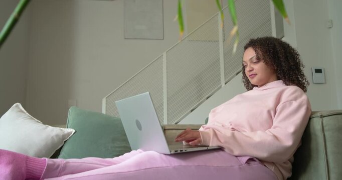 African American adult woman in pink hoodie settling onto sofa, working on laptop with mint mug