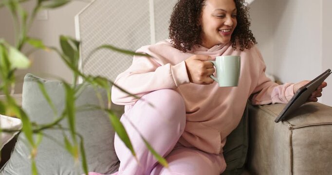 African American woman holding pale green mug, scrolling tablet and laughing while settling on sofa