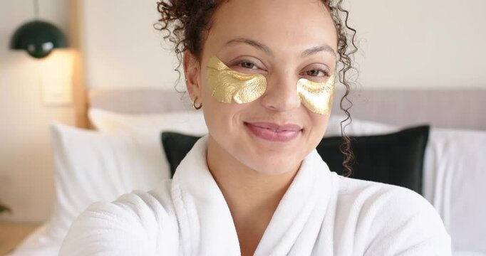 Adult African American woman opening eyes smoothing gold eye patches in robe on bed for skincare