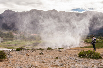 Man filming powerful geyser erupting steam in mountain landscape