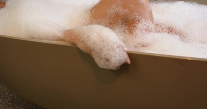 African American woman bathing in freestanding tub, adjusting forearm on tub rim with foam shifting