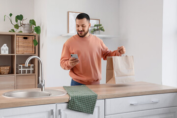 Young man with paper bag using mobile phone in kitchen. Food delivery service