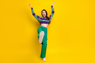 Young woman jumps with arms raised against bright yellow background wearing striped sweater and green pants for a lively fashion lifestyle stock photo.