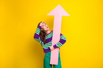Young woman in striped sweater holds a large pink arrow rising upward against a bright yellow...
