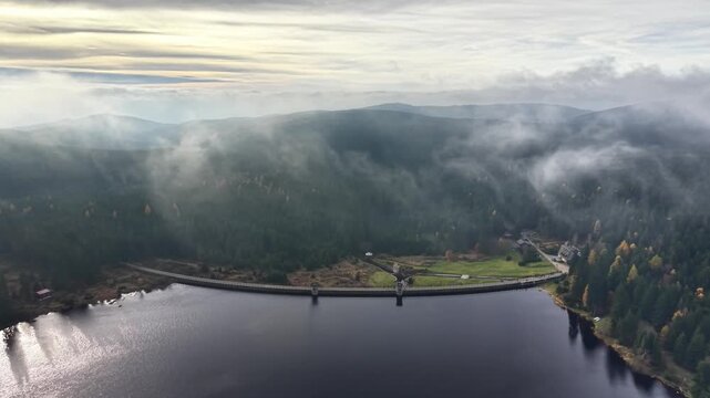 misty reservoir and dam at dawn with fog drifting over treeline, reflective water surface and subdued sunrise