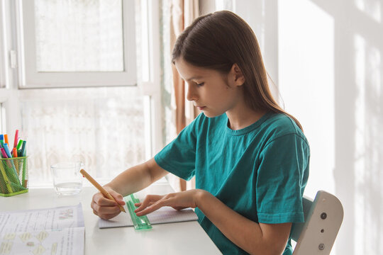 Schoolgirl studying at home at white desk near window writing in notebook with pencil and ruler during daytime, focused child doing homework in bright cozy interior with natural sunlight - Powered by Adobe