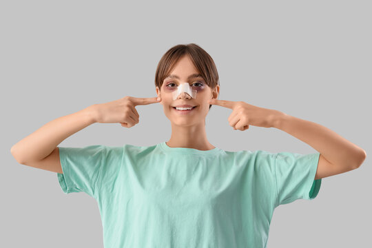 Beautiful young happy woman pointing at her bandaged nose on grey background. Plastic surgery concept