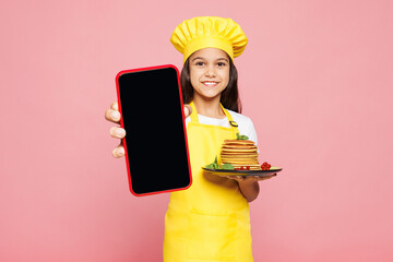 Small child girl housewife chef cook baker mother helper in yellow apron toque cap hold sweet tasty pancakes use blank screen mobile cell phone isolated on plain pink background Cooking food concept