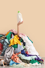 Young woman with bottle of detergent buried under pile of dirty laundry on beige background