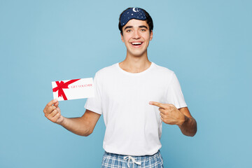 Calm young man wear pyjamas jam sleep eye mask rest relax at home hold point on gift certificate coupon voucher card for store isolated on plain light blue cyan background Good mood night nap concept