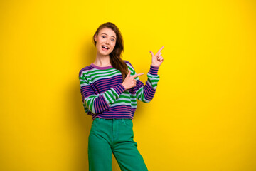 Young woman in striped multicolor sweater and green pants smiles and points against bright yellow background for lifestyle fashion stock photo