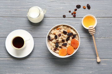 Bowl with tasty oatmeal, dried fruit, jug with milk and cup of coffee on grey wooden background