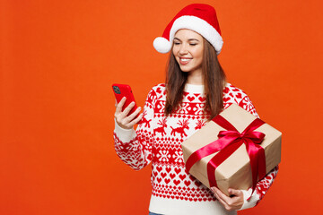 Merry young woman wear red Christmas sweater Santa hat posing hold mobile cell phone present box...