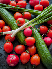 Fresh red cherry tomatoes, green leek, radish and cucumber on the black table background, top view of vegetables covered with water drops.