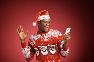 Laughing adult man wearing a festive red Christmas sweater and Santa hat while waving at a smartphone during a video call against a red studio background.