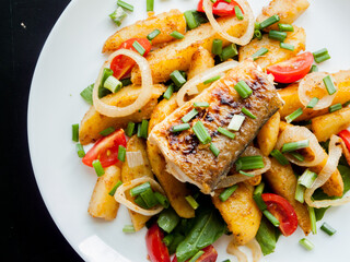 Plate with baked potatoes, piece of fish, vegetables and herbs on the table with tomato sauce.