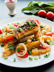 Plate with baked potatoes, piece of fish, vegetables and herbs on the table with tomato sauce.