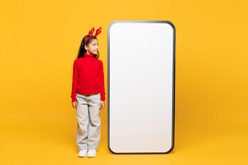 Full body merry little girl wear red clothes decorative deer horns on head posing look at big huge blank screen mobile cell phone isolated on plain yellow background. Happy New Year Christmas concept.