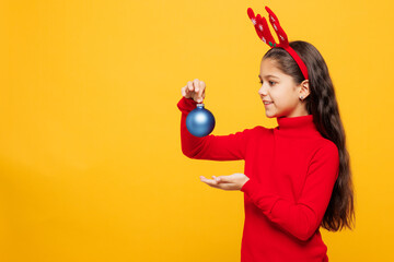 Merry little girl wearing red clothes fun decorative deer horns on head posing hold in hand ornament baubles isolated on plain yellow background. Happy New Year Christmas celebration holiday concept.