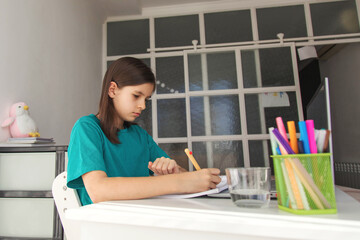 Young girl writing in notebook at a bright home desk, focusing on her schoolwork while sitting in natural daylight surrounded by stationery and study materials in a clean and organized learning space