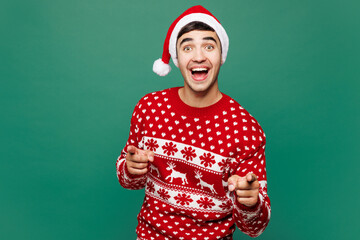Merry young man wears red warm cozy Christmas sweater Santa Claus hat posing point index finger camera on you motivating encourage isolated on plain green background. Happy New Year holiday concept.