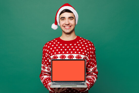 Merry young IT man wears red Christmas sweater Santa Claus hat posing hold use work blank screen on laptop pc computer isolated on plain green background. Happy New Year celebration holiday concept - Powered by Adobe