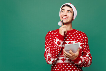 Merry young minded fun man wear red warm cozy Christmas sweater Santa Claus hat posing write down in notebook wish list isolated on plain green background. Happy New Year celebration holiday concept.