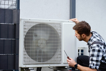 Technician performing maintenance on outdoor heat pump unit near residential building exterior. Man...