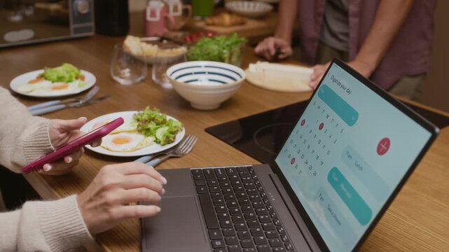 Cropped shot of couple enjoying breakfast at home, man cooking healthy meal while woman using laptop with calendar widget on screen, showing balanced morning routine