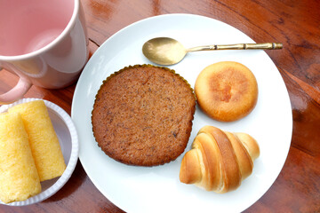 Assorted baked goods on a white plate including cake, Bakery set, Round Bun, and mini croissant with a gold spoon and pink cup, ideal for food photography and menu use, Top view