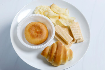 White plate with assorted snacks including a croissant, filled bun, potato chips, and wafer sticks. Clean and simple setup suitable for food menus, Top view