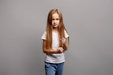 Aggressive young child girl clenching fists in emotional defensive pose, expressing frustration and defiance in studio portrait on gray background, showing intense feelings and reaction