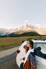 Naklejka premium Woman car mountains road travel landscape: young woman leans on a white SUV beside a mountain road, cozy sweater and soft sunlight over valley and peaks during an outdoor road trip.