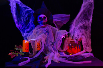 Halloween decoration with a skeleton wearing a witch hat, surrounded by spiderwebs, pumpkins, and candles, illuminated by purple and red lighting.