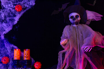 Halloween decoration: a witch's skeleton on a black background, surrounded by cobwebs, pumpkins, and candles illuminated with purple and green lights. A card backdrop.