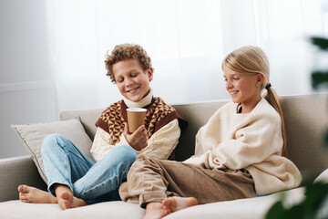 Two children sit on a cozy sofa, sharing a warm moment with mugs in hand, dressed in soft sweaters and jeans, bathed in bright natural daylight and gentle home ambience.