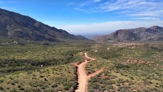 Aerial view of Four Peaks Wilderness showcasing a winding dirt road amid green vegetation against a backdrop of mountains and a clear blue sky, Apache Junction, Arizona, United States.
