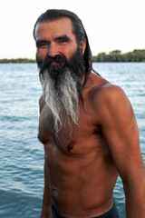 Bearded shirtless man standing in river with crossed arms at sunset