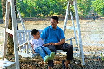 A father and his young son sitting and bonding on an outdoor swing near a natural landscape.