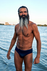 Bearded shirtless man standing in river with crossed arms at sunset