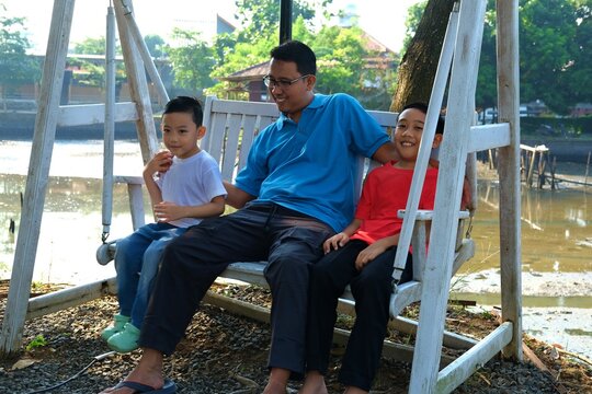 A father relaxing with his two children on a swing at an outdoor recreational area. - Powered by Adobe
