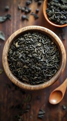 Dried green tea leaves in wooden bowl on rustic table with scattered herbs and spoon for healthy beverage preparation and culinary cooking.