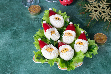 Festive deviled eggs in the shape of Santa Claus on lettuce leaves on Christmas tree-shaped plate on green background