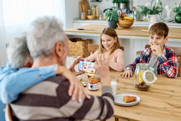 Portret of brother and sister, grandchildren and grandparents having fun together taking a selfie photo with a mobile phone and eating breakfast in kitchen