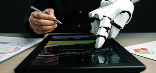 A robot hand interacts with digital data on a tablet screen while a human hand writes notes on business charts, representing collaboration between technology and human efforts.Noogenesis