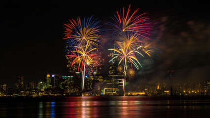 Fototapeta premium Spectacular nighttime fireworks display illuminates a modern city skyline reflecting vibrantly on the water below New Year’s Eve