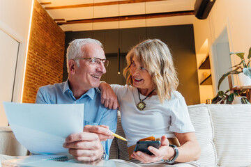 Elderly couple happily managing household finances and planning future retirement, reviewing important documents and bills together in their comfortable home