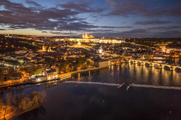 Fototapeta premium An aerial view of Prague at night. The Vltava River flows beneath the Charles Bridge while the castle is illuminated in the distance. Buildings and streets shine with lights.