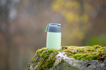 Thermos flask with hot coffee or tea drink is standing on a moss covered rock in a forest, traveling and wanderlust concept, food container