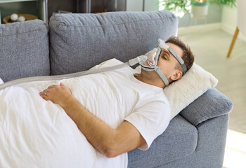 Obraz premium Man wearing oxygen mask sleeping on sofa. Patient uses CPAP therapy at home on a pillow. Tubing connects to a device, suggesting healthcare support and recovery. Concept: home respiratory care.